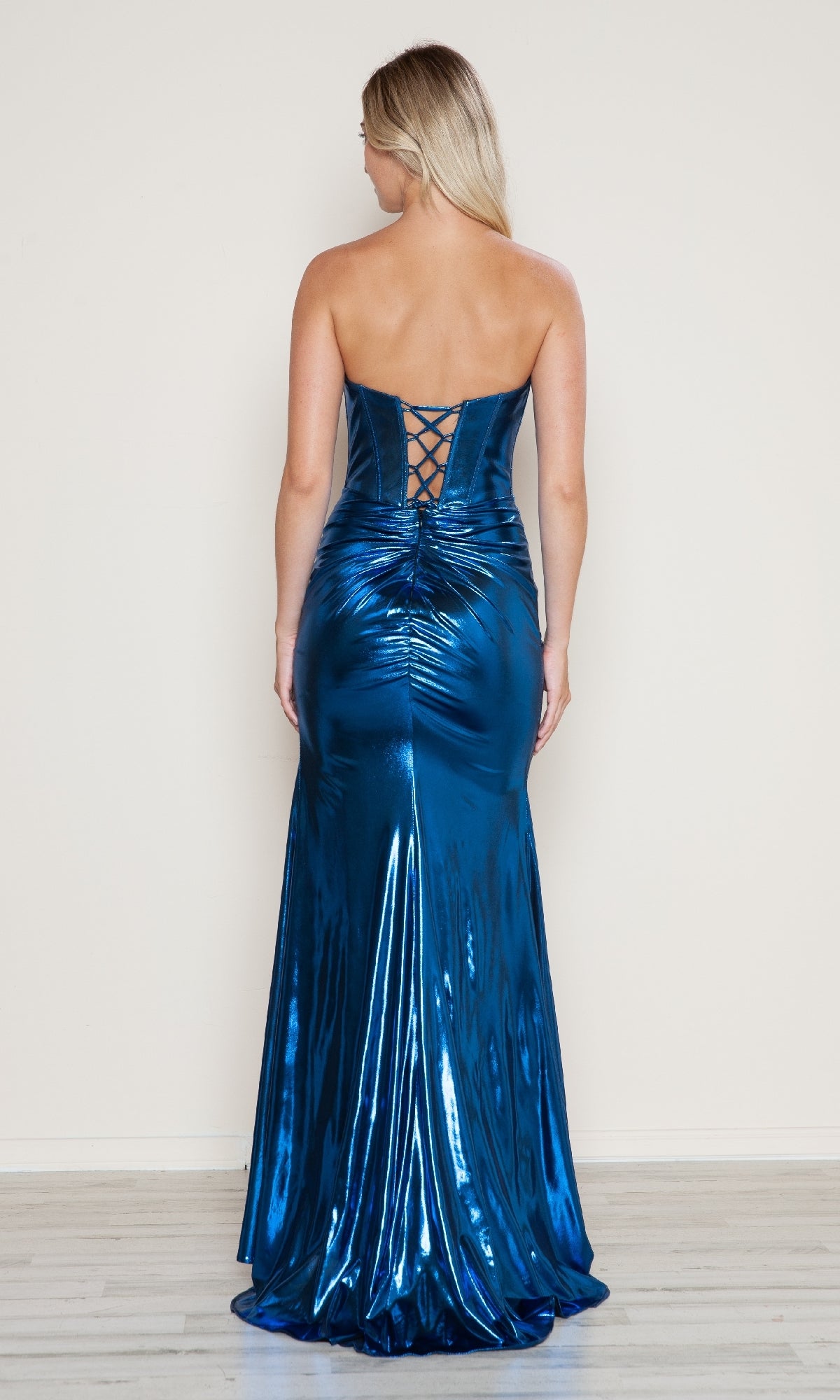 A woman faces away from the camera, modeling the Poly USA 9684 Lace-Up Strapless Long Prom Gown—a shiny blue metallic dress with a slight train—against a plain background.